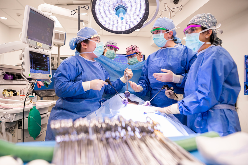 a group of eight people with a face shield, blue medical mask, and blue scrubs stand in a medical office room with desks